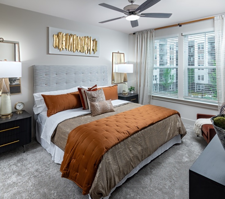 A bedroom in a Duluth, GA apartment for rent at Elan Satellite Place with a large bed with decorative pillows and sheets, two black side tables with lamps and wall mirrors, a front-end table with decorative home decor pieces, a chair with a blanket on it,