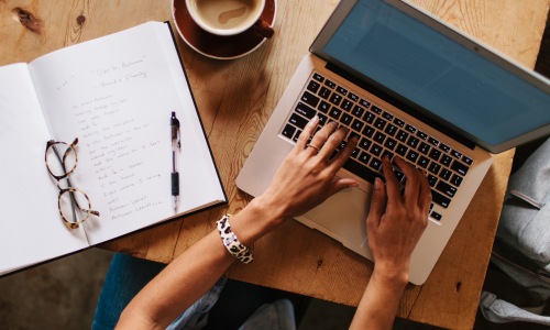 womans arms typing on laptop notebook open glasses coffee cup pen